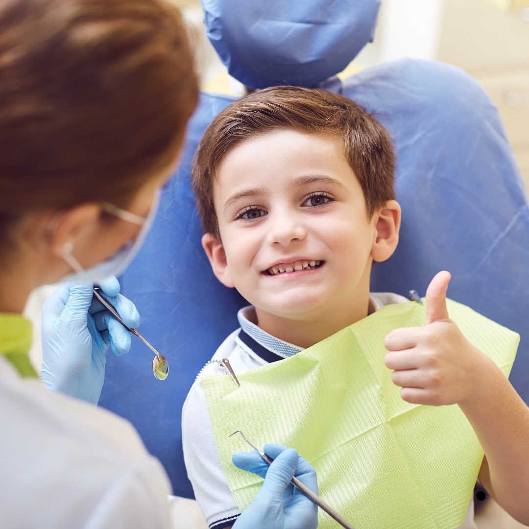 Menino sorrindo e fazendo sinal de positivo na cadeira do dentista durante um atendimento acolhedor